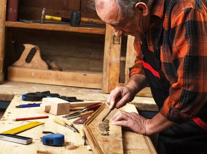 A woodworker in a flannel shirt carves a wooden piece on a workbench filled with various tools.