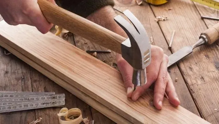 A person uses a hammer to drive a nail into a wooden board on a workbench with woodworking tools nearby.