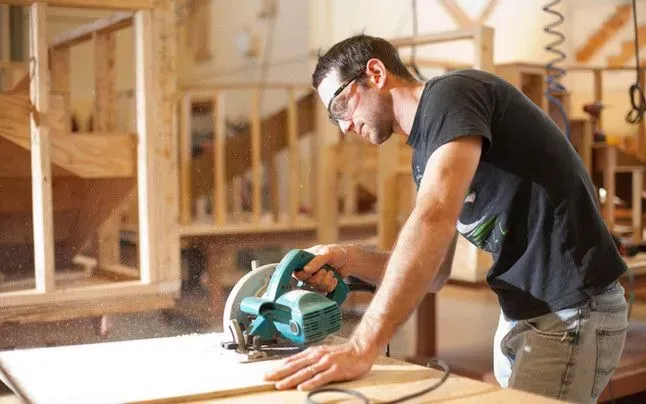 A carpenter wearing safety goggles uses a circular saw to cut a piece of wood in a workshop.