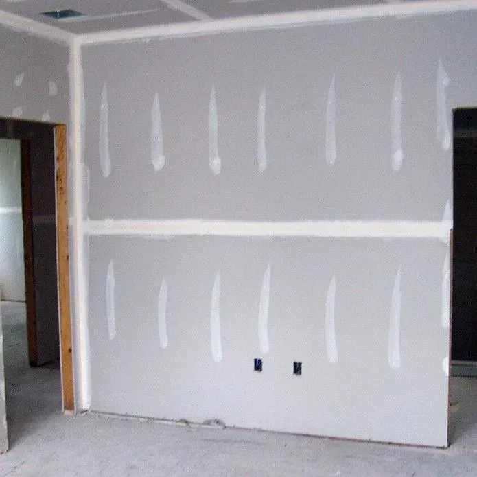 A grey drywall wall in a room under construction, with taped seams over joints and screw heads, and two electrical outlets.