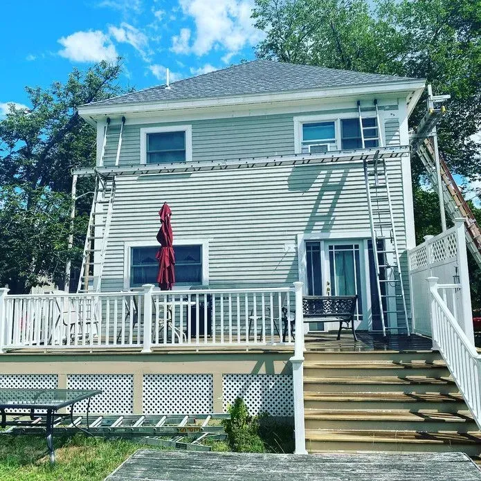 A two-story light gray house with white trim, a large deck, and metal ladders propped against the exterior.