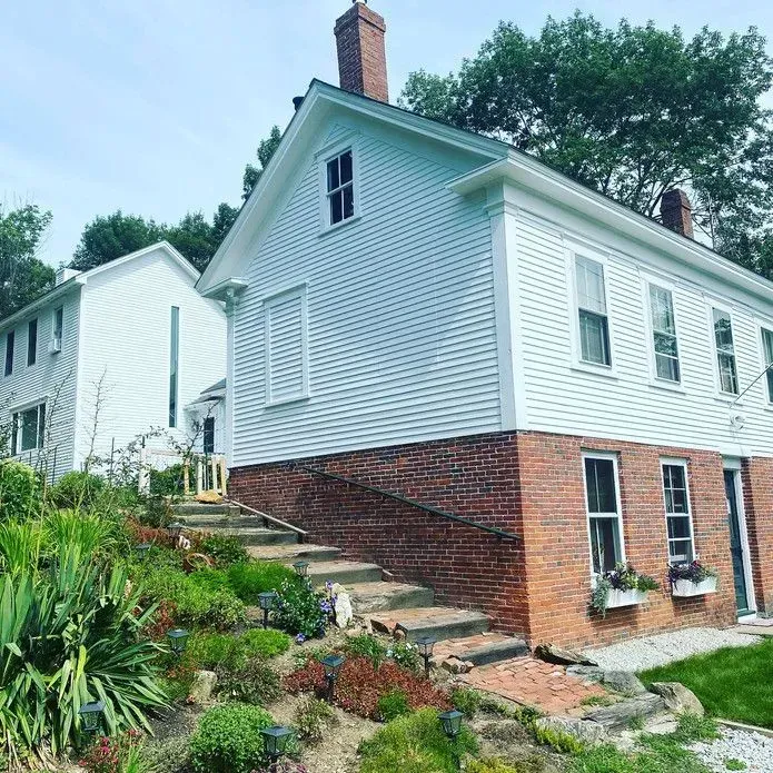 A light blue house with a brick foundation and a brick chimney, featuring stairs leading up the landscaped yard.