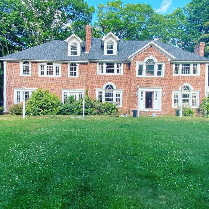 A large, two-story brick colonial-style home with multiple windows, a gabled roof, and green lawn under a sunny blue sky.