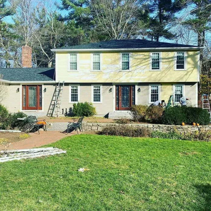 A two-story, yellow colonial-style house sits in a sunny yard with green grass, surrounded by tall pine trees.