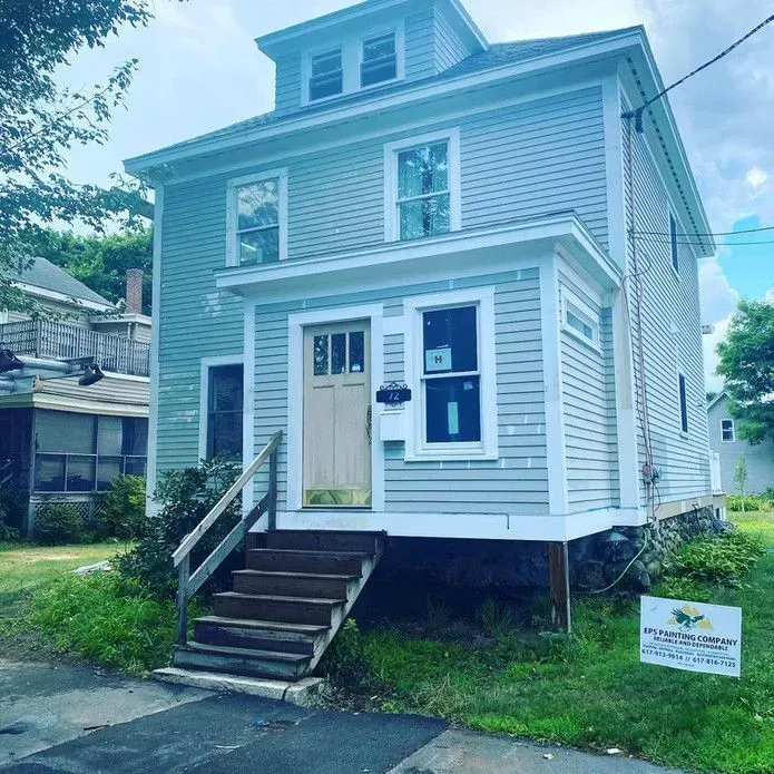 A light blue two-story house with a white front porch, a tan door, and stairs leading to the entrance on a sunny day.