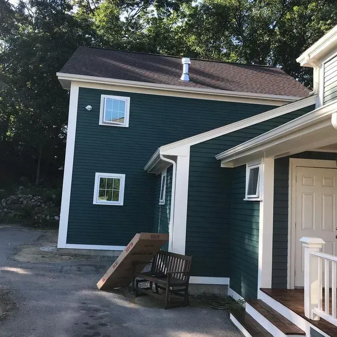 A house with dark teal siding and white trim, featuring a wooden bench and a large cardboard box leaning against the wall.