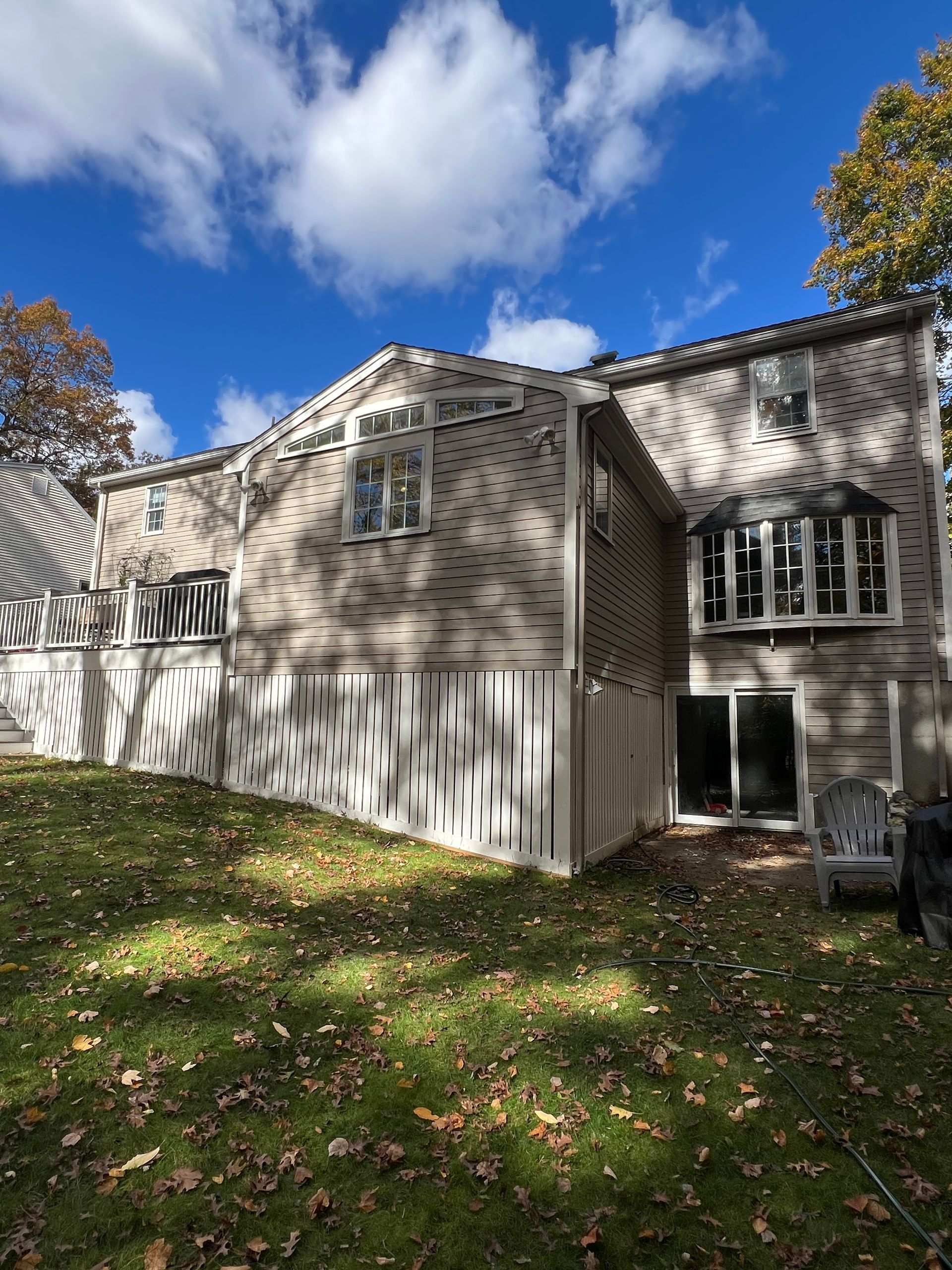 A two-story house with light-colored brick, white trim, and a white deck, viewed from a yard covered in autumn leaves.