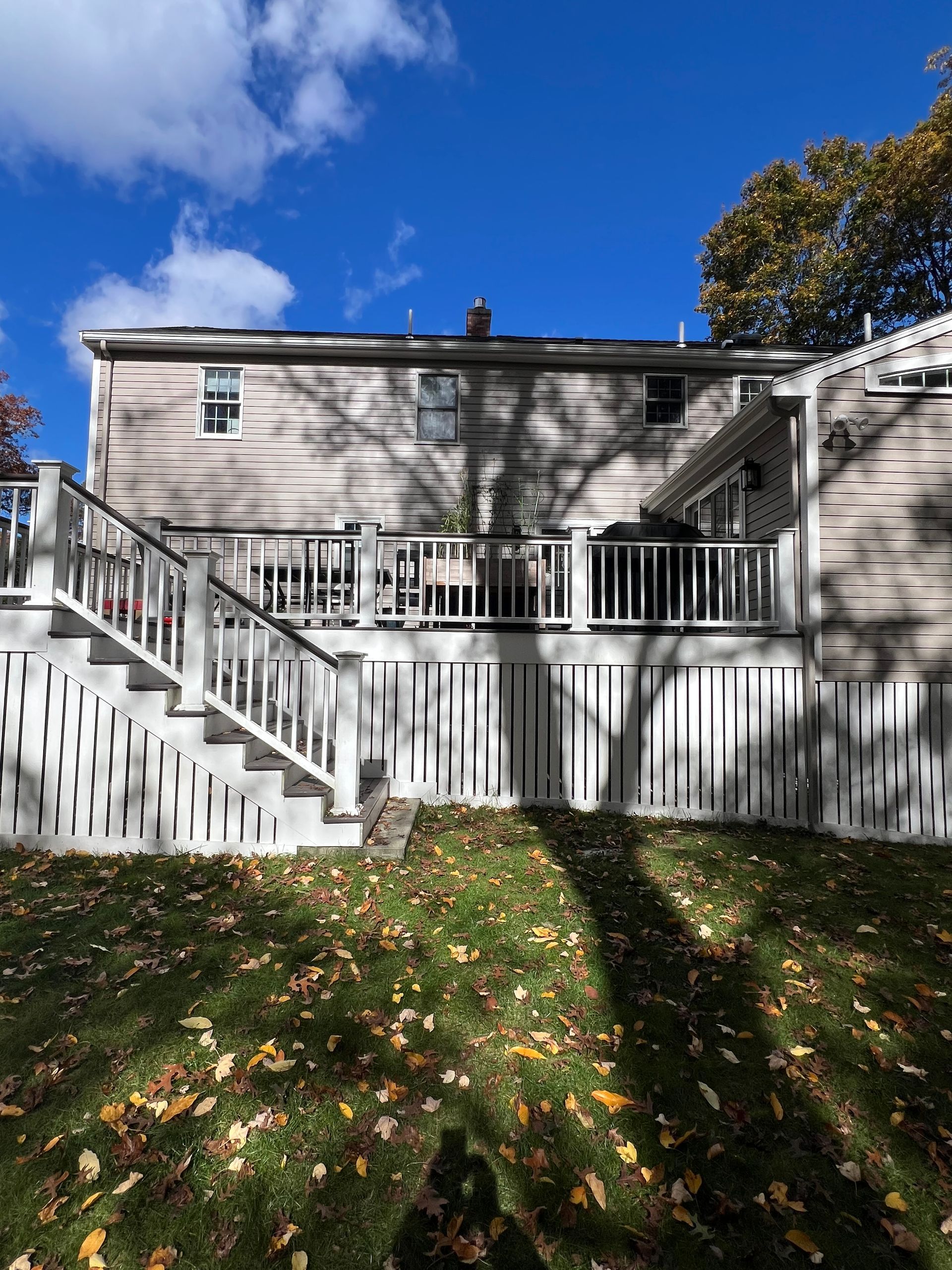 A light-colored house with a large wooden deck, stairs, and white lattice skirting, set in a yard covered in autumn leaves.
