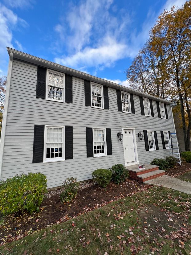 A two-story grey colonial home with black shutters, white trim, and a front door surrounded by autumn trees under a blue sky.