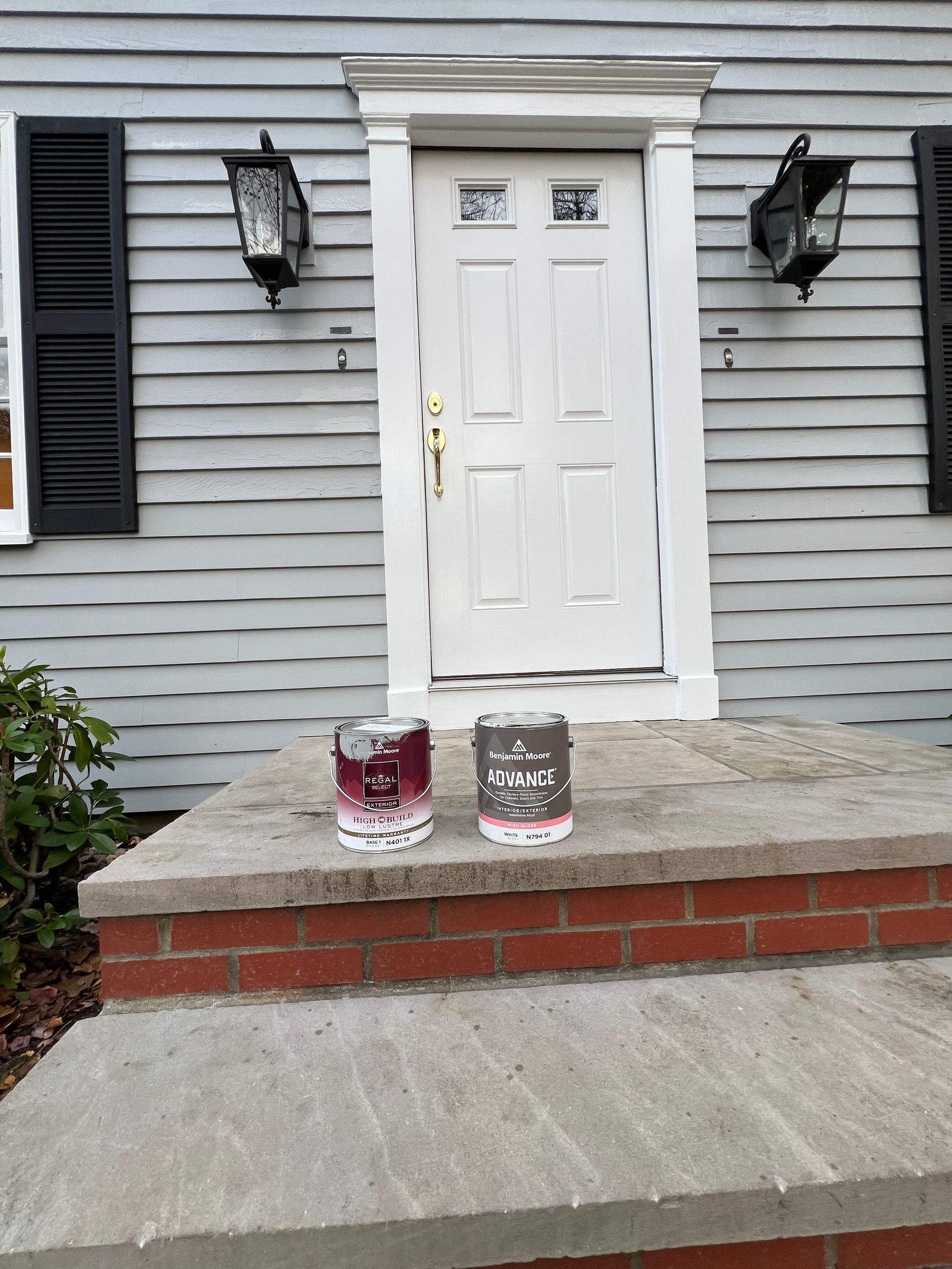 Two paint cans, one dark red and one grey, sit on the brick and stone front steps of a house with a white door.