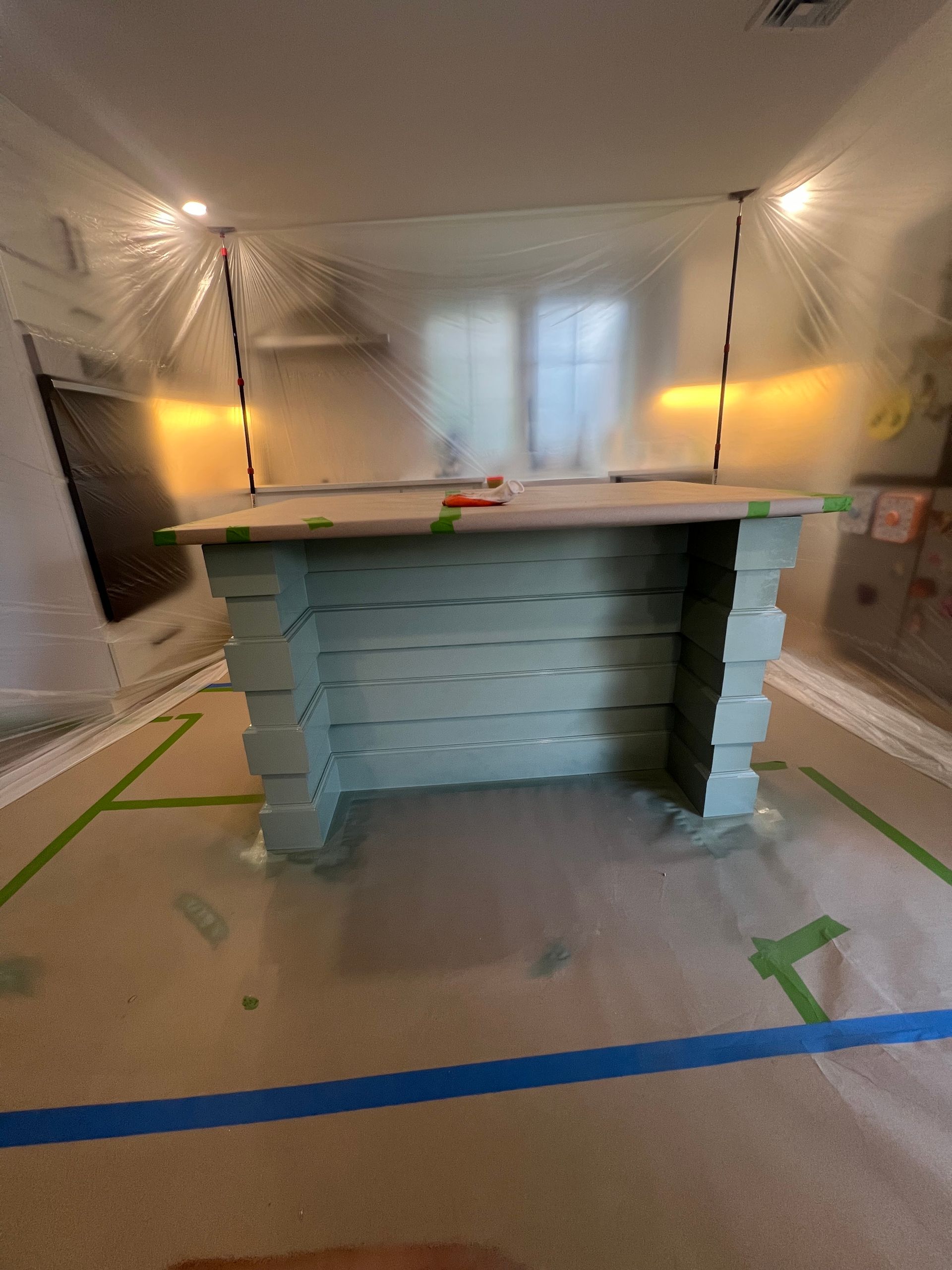 A kitchen island painted soft green, surrounded by plastic protective sheeting and painter's tape on the floor.