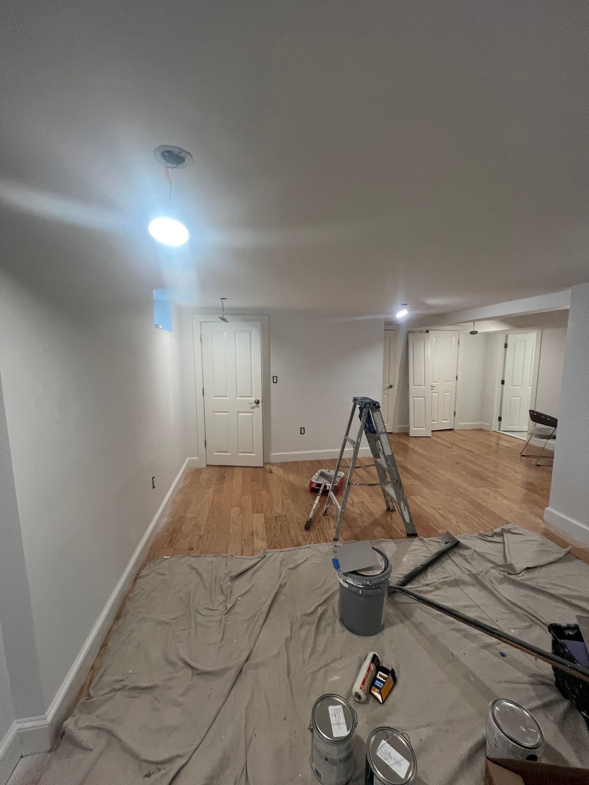 An interior room under renovation with light-colored walls, hardwood floors, a ladder, paint supplies, and drop cloths.
