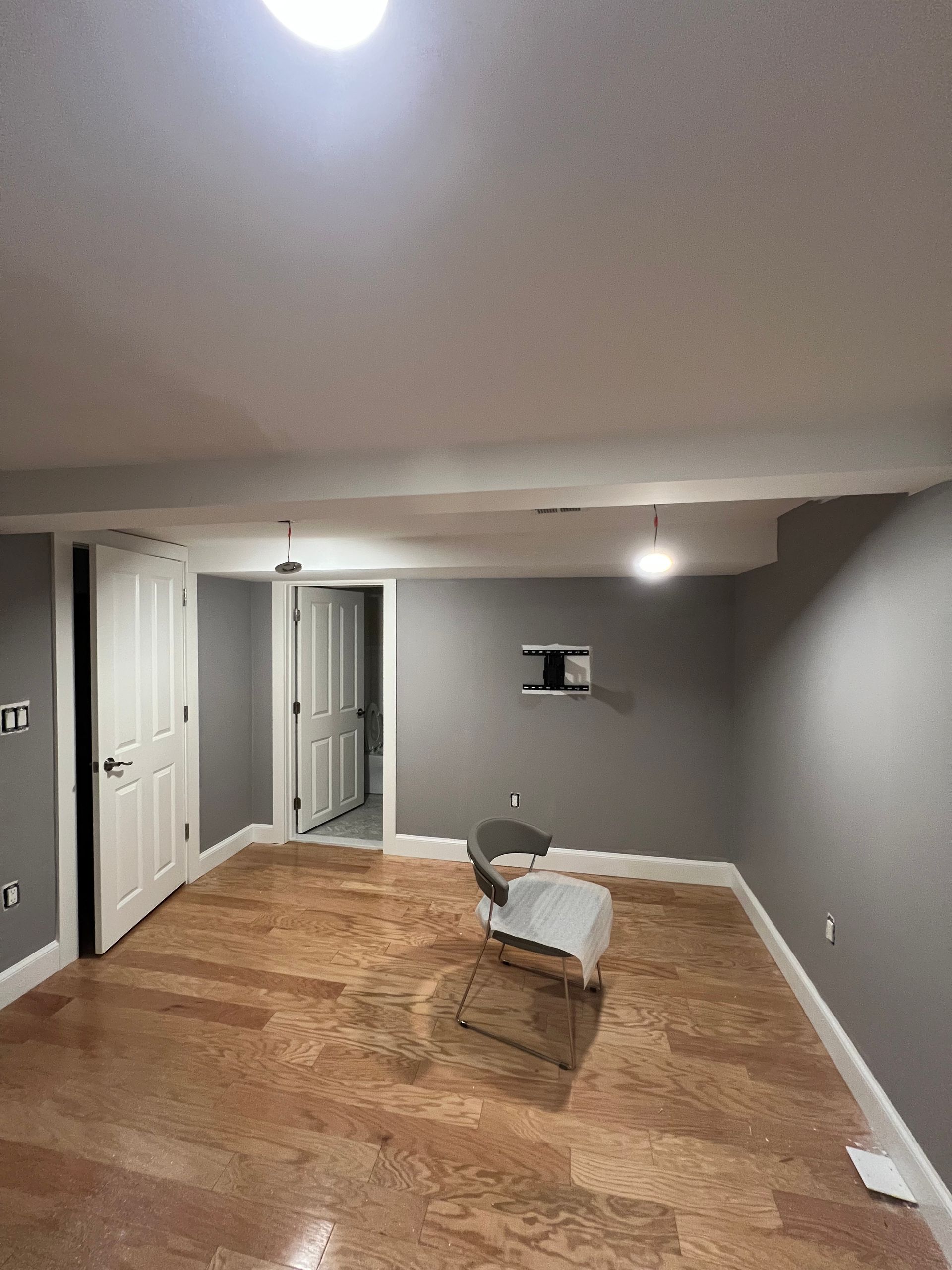 A vacant room with gray walls, wood-grain flooring, two white doors, and a single chair sitting in the middle of the space.