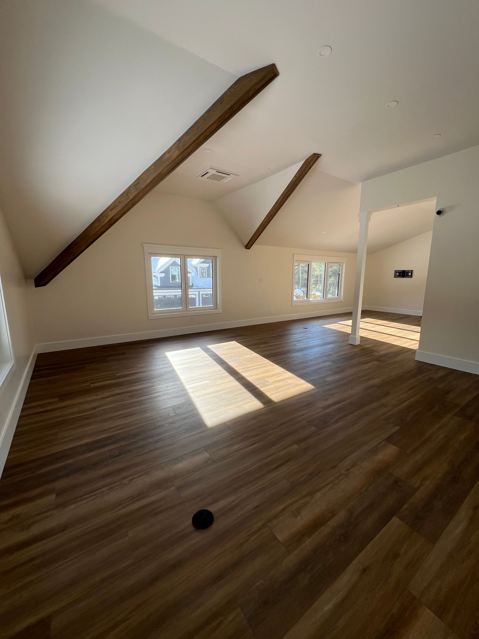 An empty attic room with angled ceilings, exposed wooden beams, brown wood-look flooring, and sunlight hitting the floor.