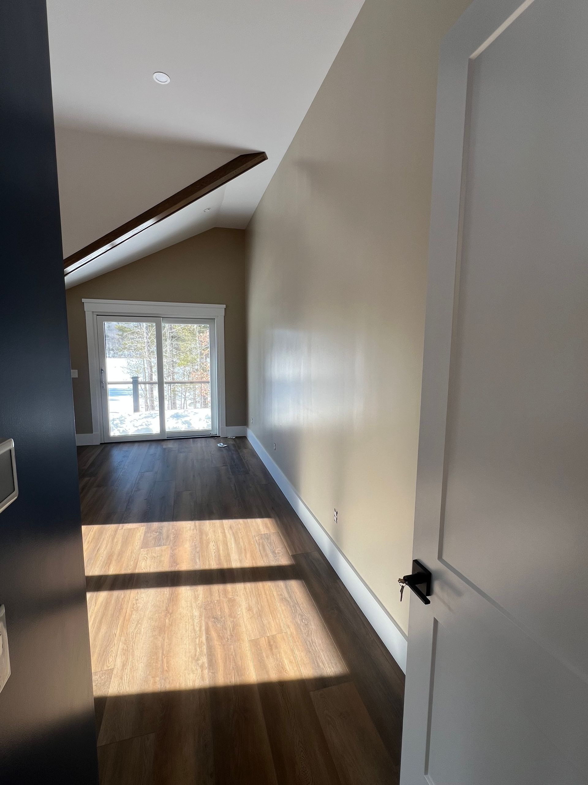View from a doorway into a long, sunlit room with wood-look floors, beige walls, and glass sliding doors.
