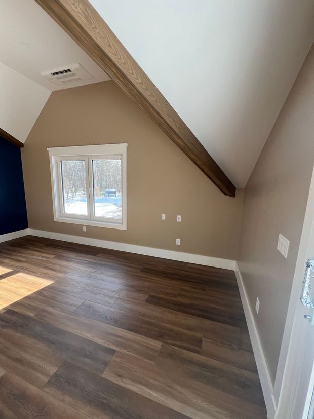 An empty room with tan walls, a slanted ceiling with a wooden beam, dark wood floors, and a window overlooking snow.