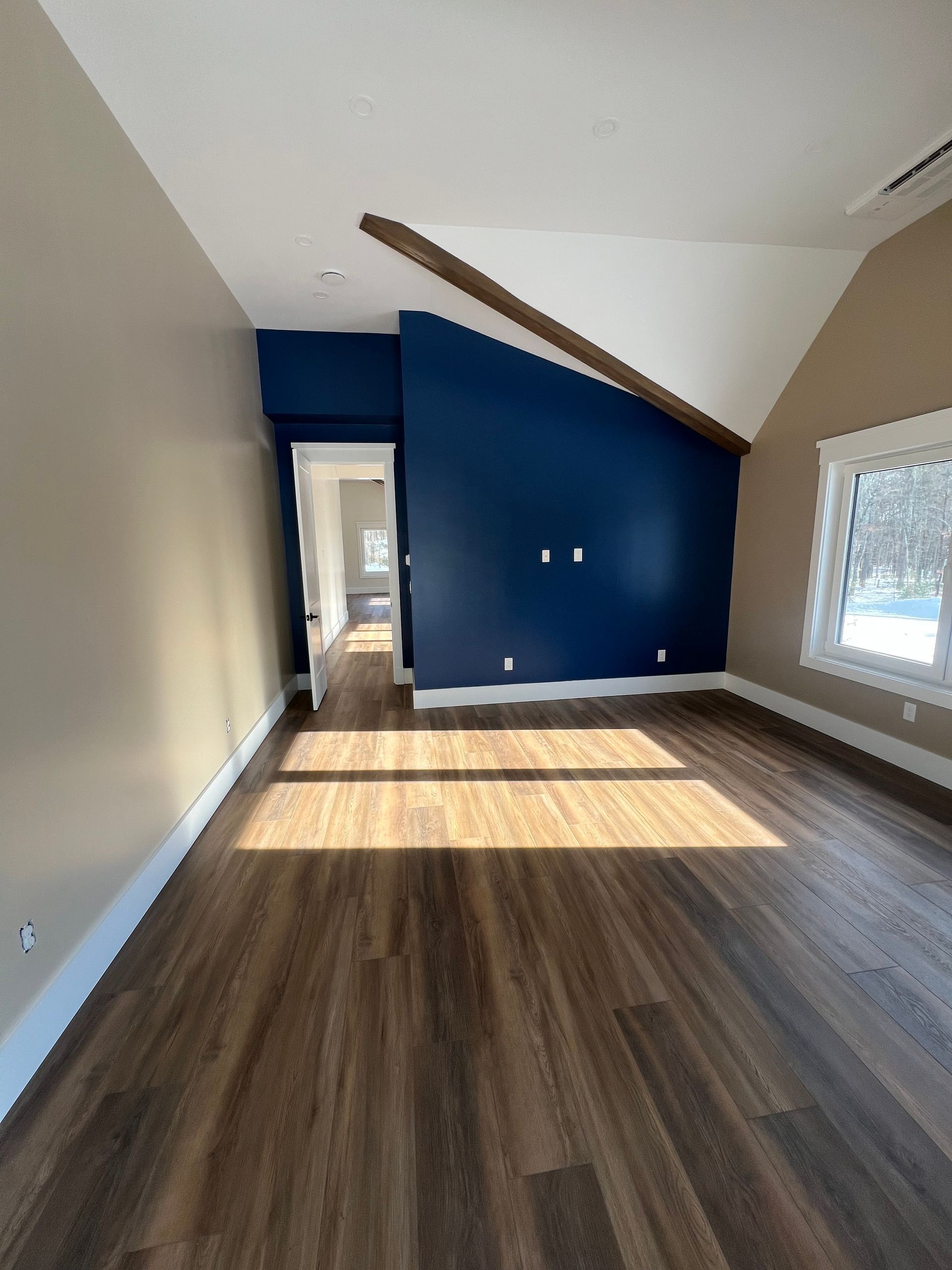 An empty room with wood-look flooring, beige walls, and a blue accent wall under a sloped ceiling with a wood beam.