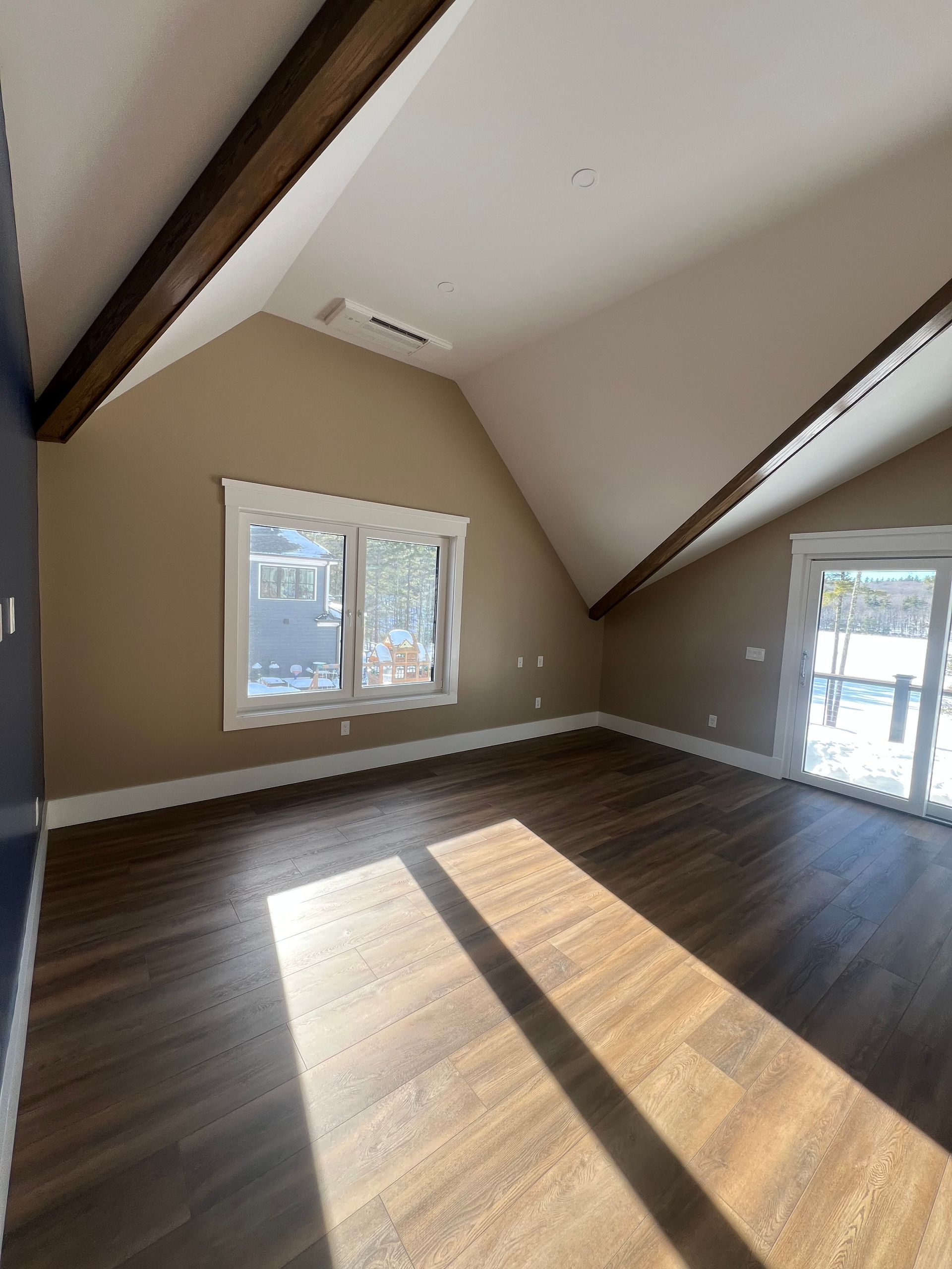 An empty room with vaulted ceilings, dark wood-look floors, tan walls, a window, sliding glass doors, and exposed beams.