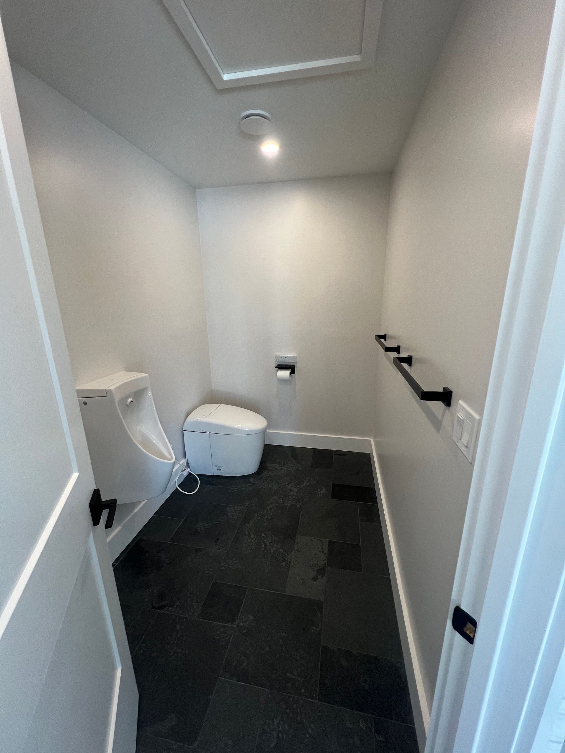 A small restroom with a white urinal, a white toilet, black tiled flooring, and a black towel bar on the right wall.