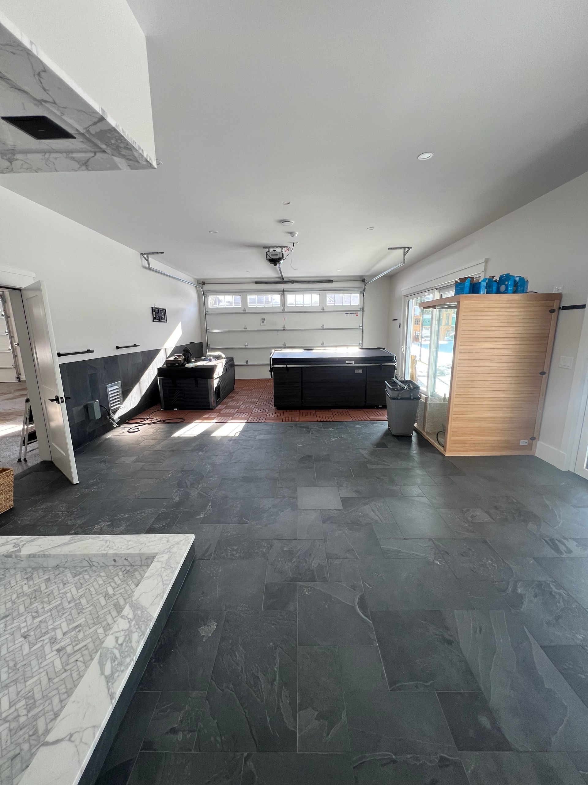 A wide garage with dark slate-style flooring, a workbench, a sauna, and an open garage door letting in daylight.