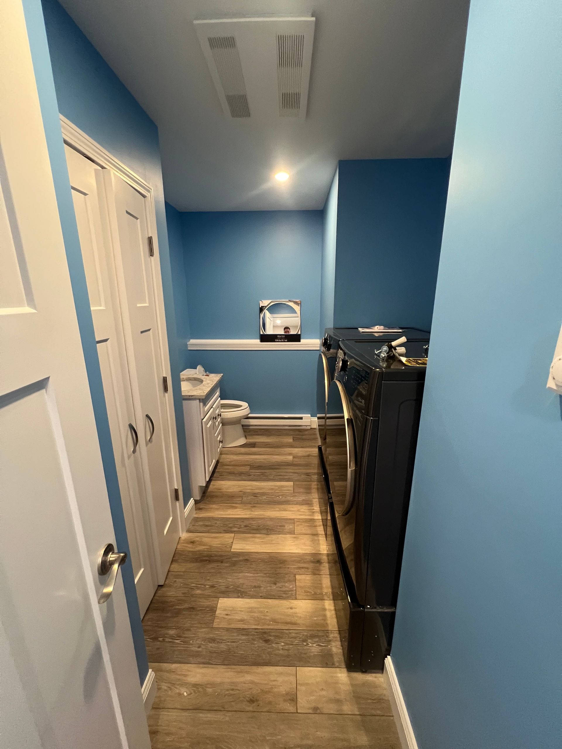 A narrow laundry room with blue walls, wood-look flooring, a white vanity, a toilet, and a dark side-loading dryer.