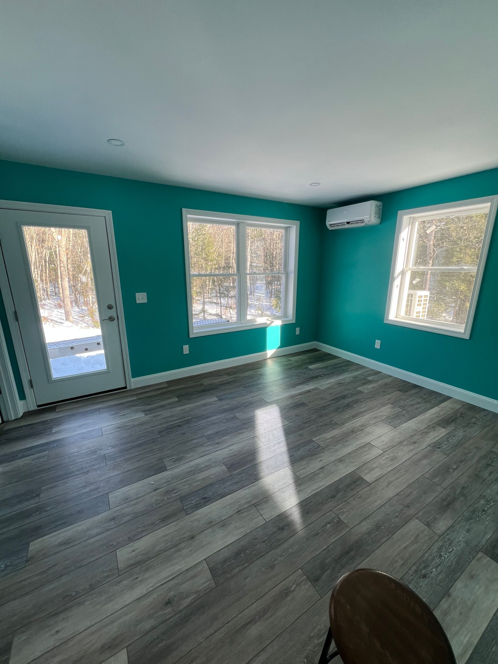 A room with teal walls, wood-look flooring, two windows, a glass-pane door, and a wall-mounted mini-split AC unit.