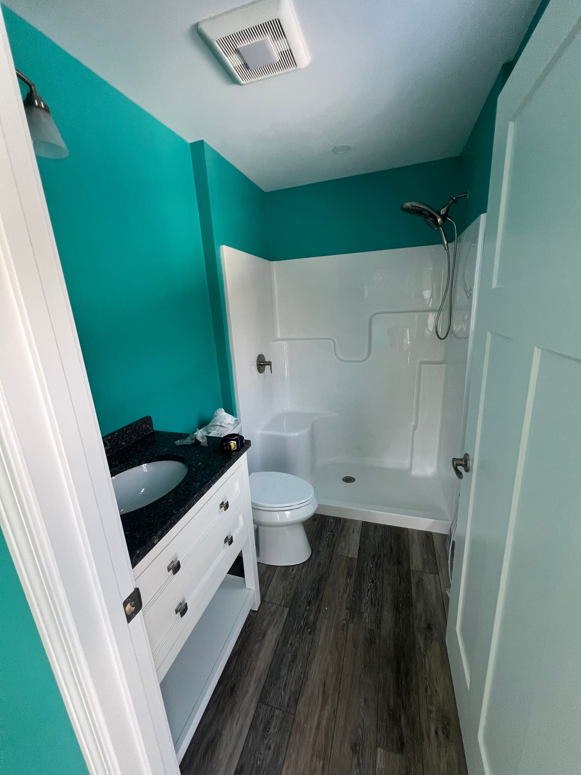 A small bathroom with teal walls, white vanity, dark countertop, toilet, and a white walk-in shower stall with wood floors.