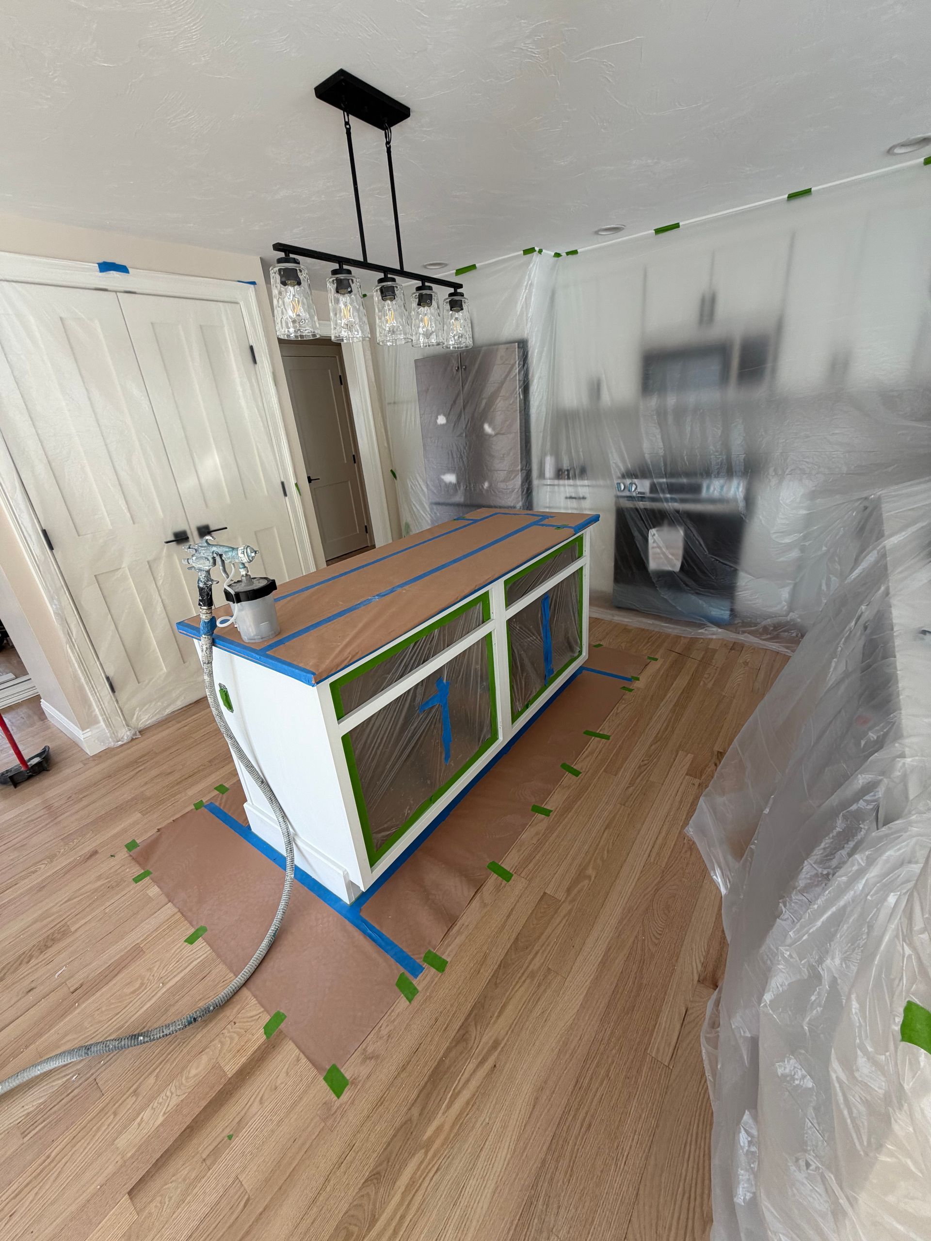 A kitchen island is taped off and being prepped for painting, with a spray gun sitting on top and plastic sheeting nearby.