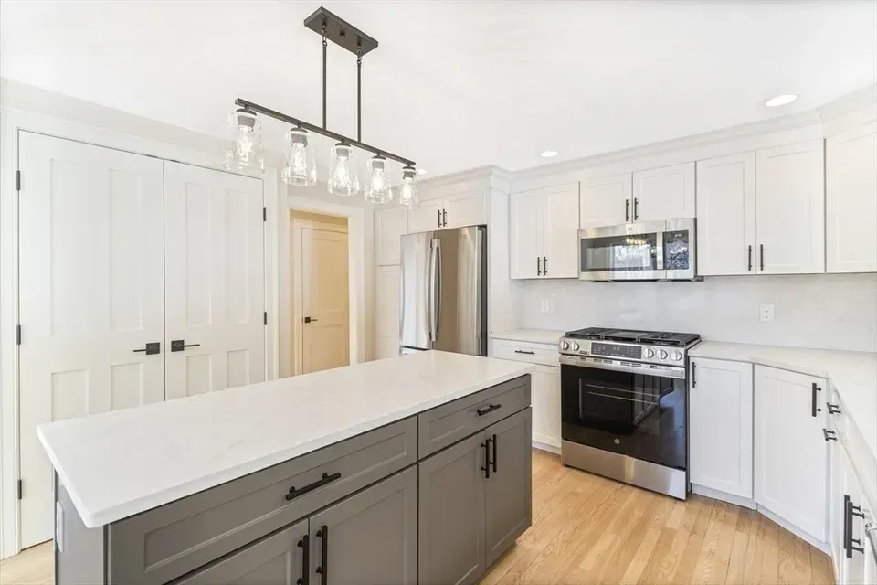 Modern kitchen with white upper cabinets, a gray central island, stainless steel appliances, and light hardwood floors.