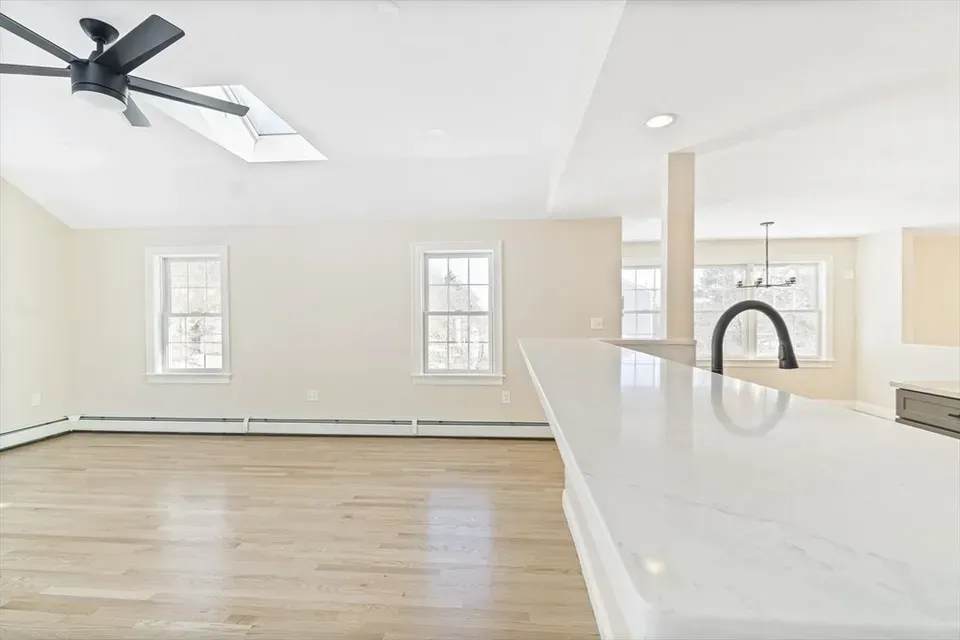 An open-concept room with a light-colored wood floor, white walls, a ceiling fan, and a view into a kitchen area.
