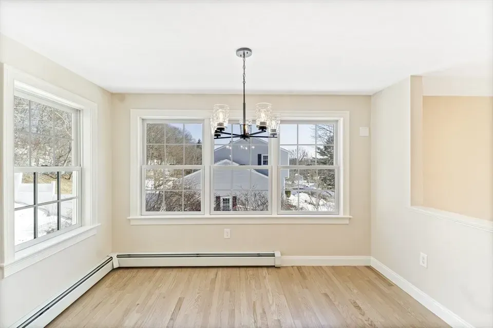 An empty dining room with light hardwood floors, beige walls, windows with white trim, and a black metal chandelier.