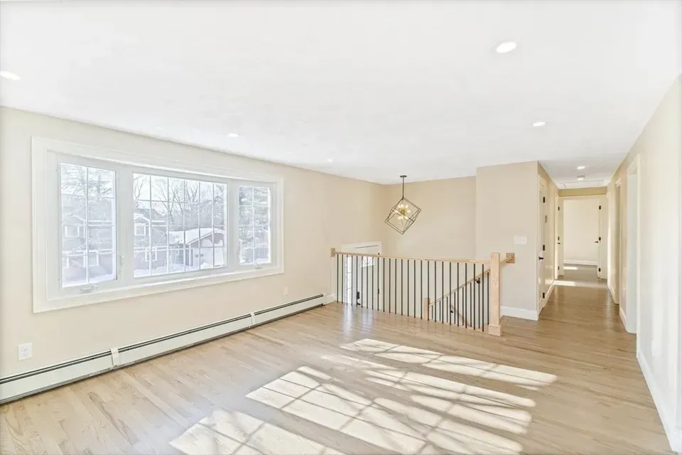 A bright, empty room with light wood floors, a large window, and a staircase with vertical railings.