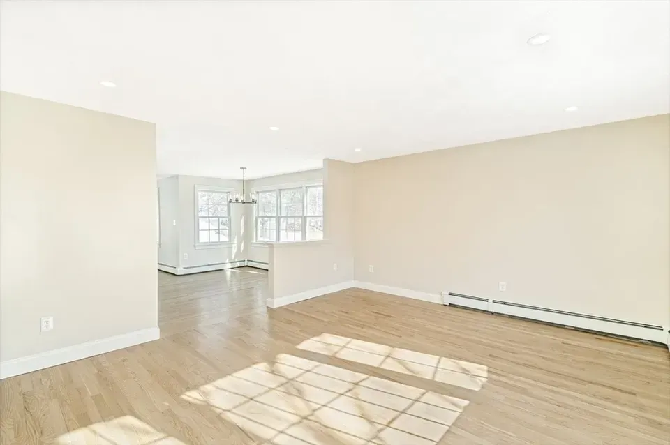 A bright, empty living room with light beige walls, light wood flooring, recessed lighting, and large windows.