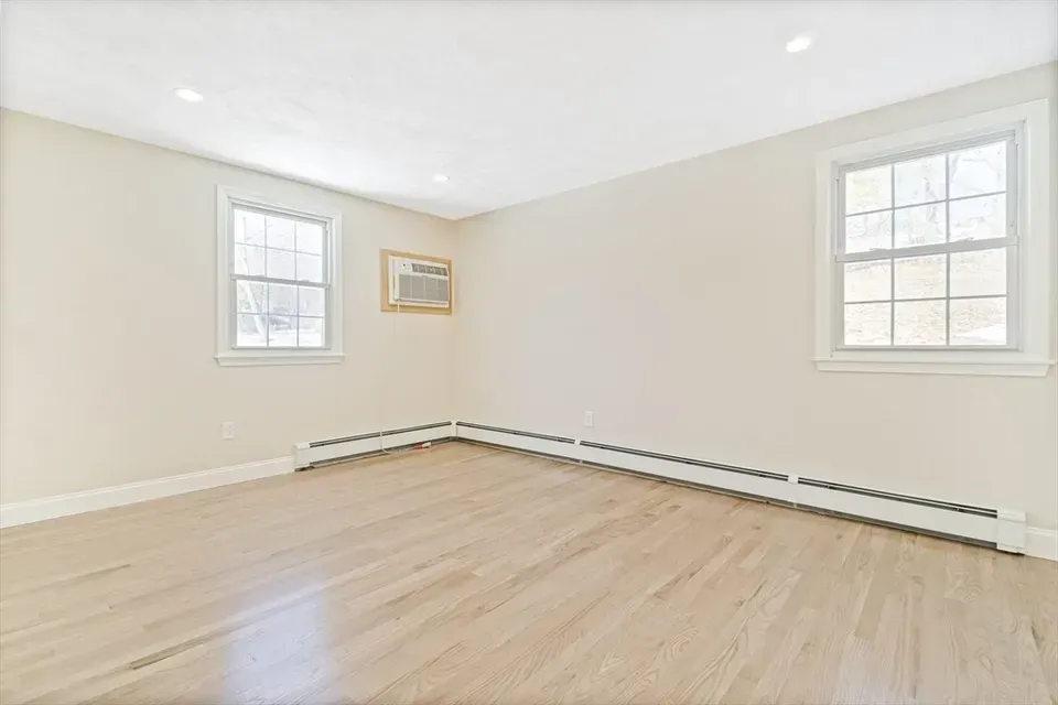 An empty room with light hardwood floors, beige walls, two windows, and white baseboard heating.