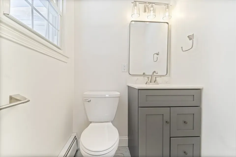 A half bathroom with white walls, a white toilet, and a grey vanity cabinet with a rectangular mirror and light fixture.