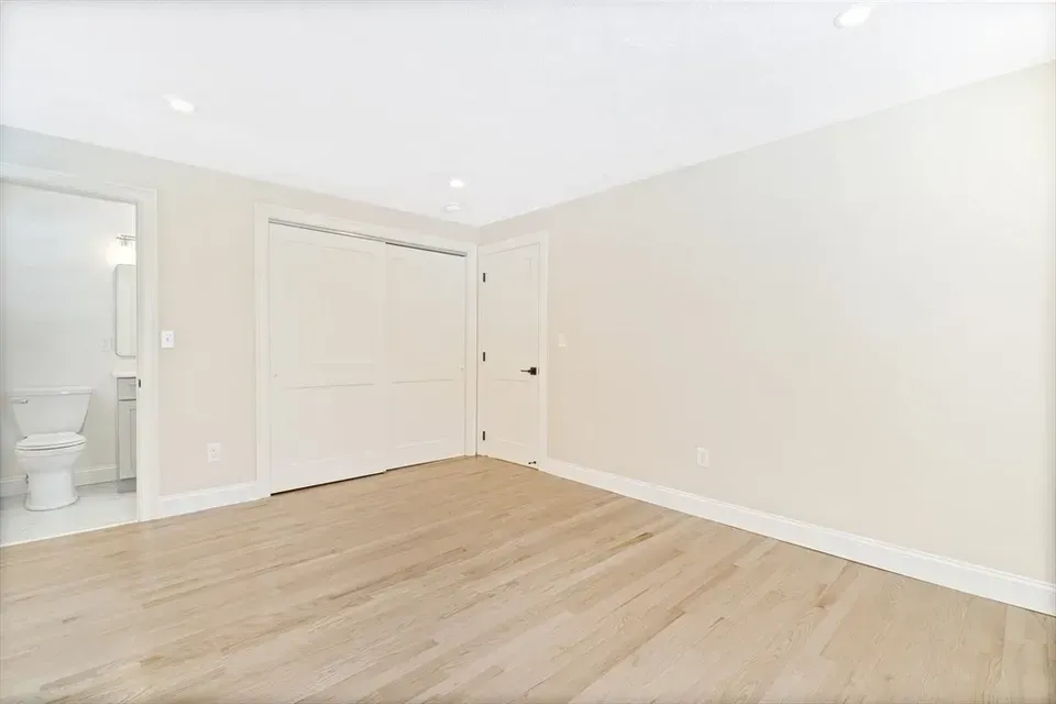 A bright, minimalist room with pale wood floors, off-white walls, a closed closet, a standard door, and an attached bathroom.