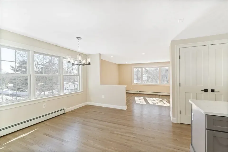 A bright dining area with light wood floors, a chandelier, and an open layout leading to a living space with large windows.