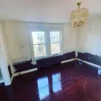 Empty room with dark hardwood floors, white walls, and a large window, under a decorative gold chandelier.