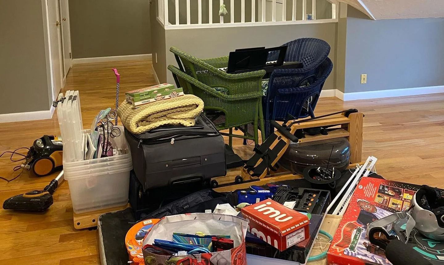 A room filled with various household items, moving supplies, chairs, and boxes scattered across a wooden floor.