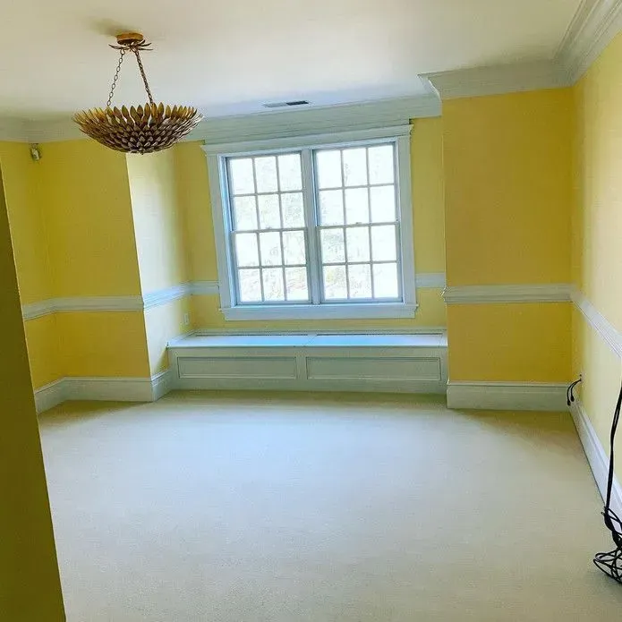 An empty room with bright yellow walls, white trim, beige carpeting, a window bench, and a modern gold chandelier.