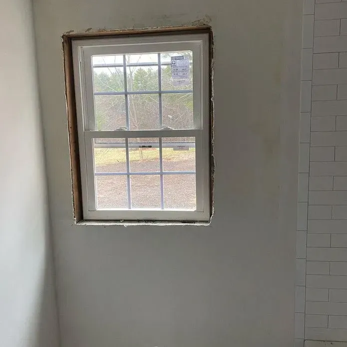 A white-framed window with a grid pattern looks out onto trees, set into a plain wall next to a white tiled surface.