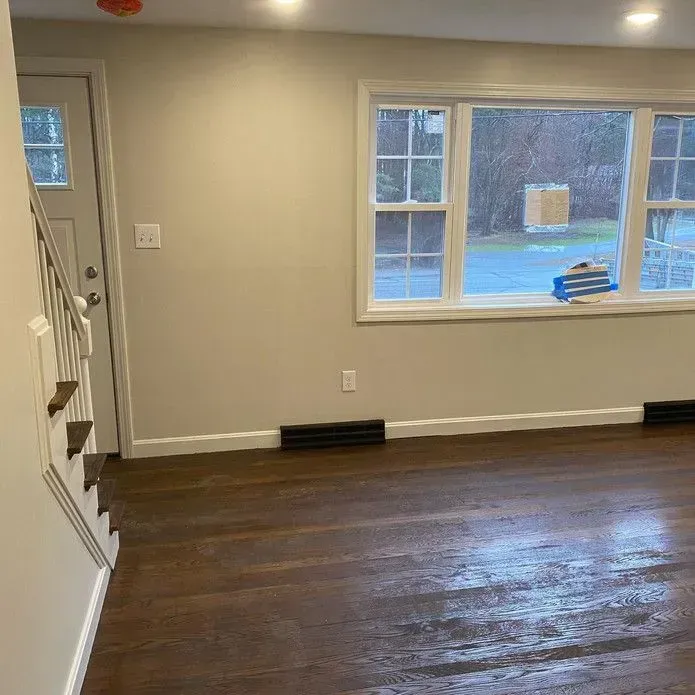 An interior room featuring dark hardwood floors, cream walls, a white door, and a large window looking out onto a yard.