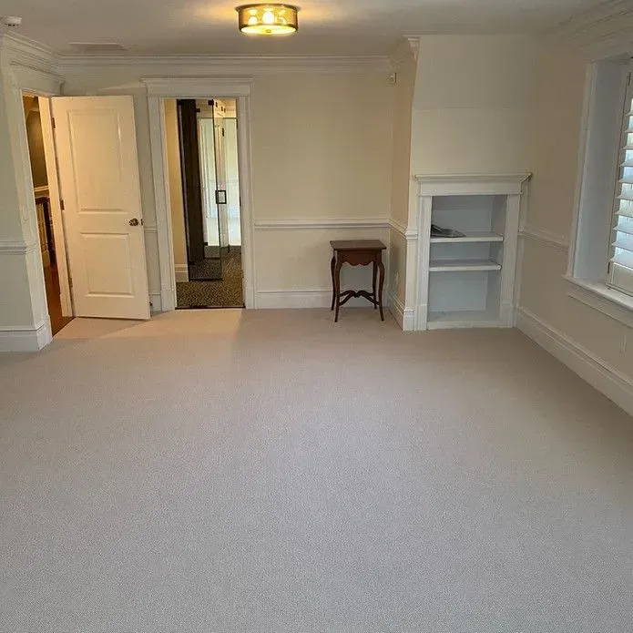 An empty room with light carpet, white walls, a doorway to another room, a small wooden side table, and a built-in shelf.