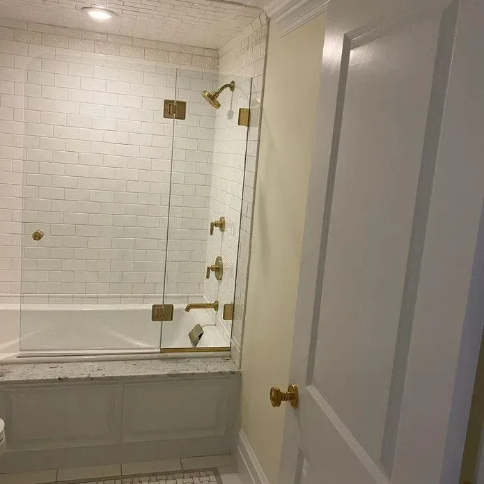 A modern white bathroom with a tiled bathtub area, glass shower enclosure, and gold fixtures next to a white door.
