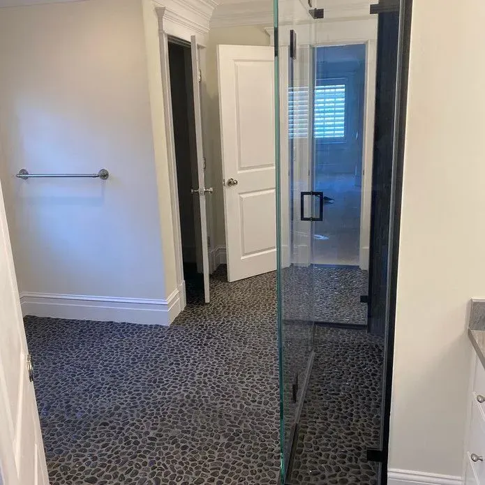 A bright bathroom features a walk-in shower with a glass door, pebble-patterned flooring, and white paneled doors.