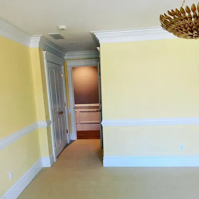 A room with yellow walls, white trim and crown molding, a tan carpeted floor, and a doorway leading into a hallway.