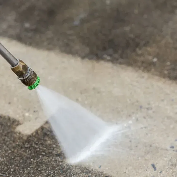 A pressure washer nozzle sprays a fan of water onto a dirty concrete surface, cleaning a clean white path.