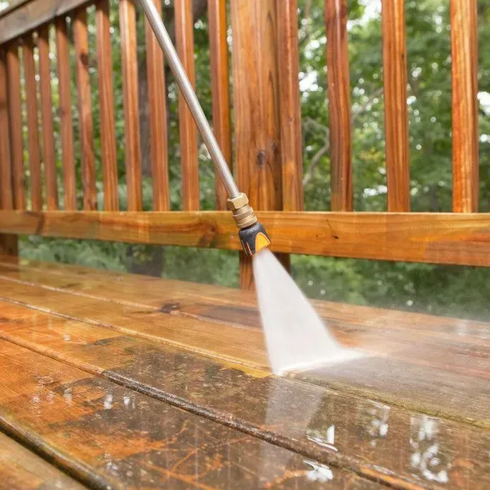 A pressure washer sprays a high-pressure stream of water onto a wooden deck, cleaning a patch of dirty surface.