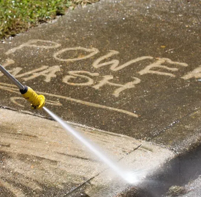 A pressure washer wand sprays a concrete sidewalk, revealing clean pavement underneath the text 