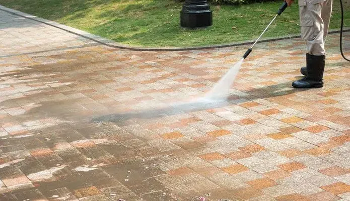 A person in boots pressure-washes a dirty brick walkway, revealing clean tiles underneath.